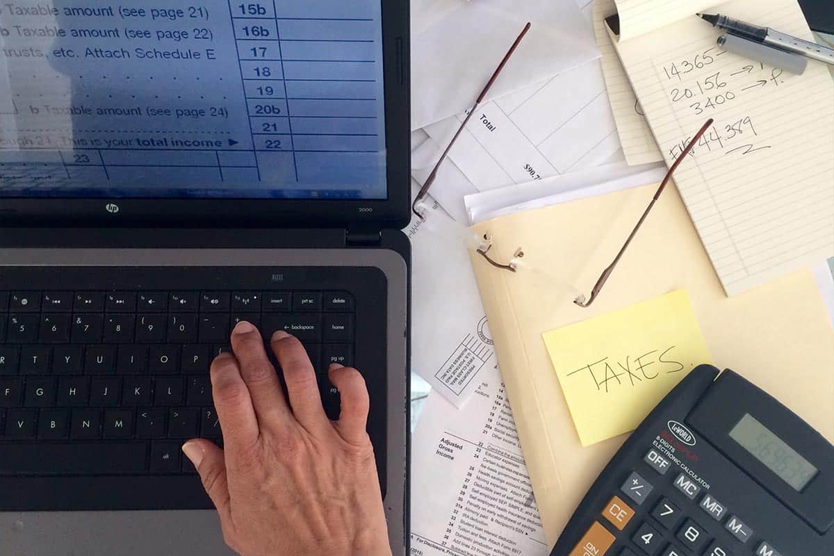 Man using laptop to compute personal income tax relief figures on a desk full of documents, a folder labelled taxes, eyeglasses and a black calculator